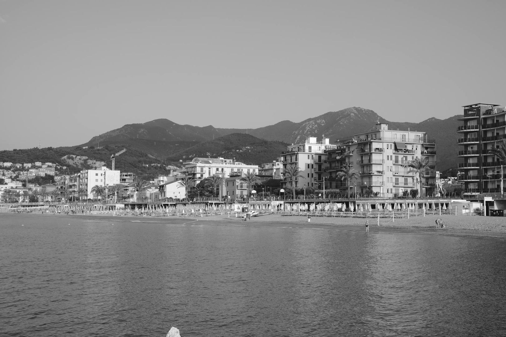 Svartvit bild av strand, hav och byggnader med berg i bakgrunden.